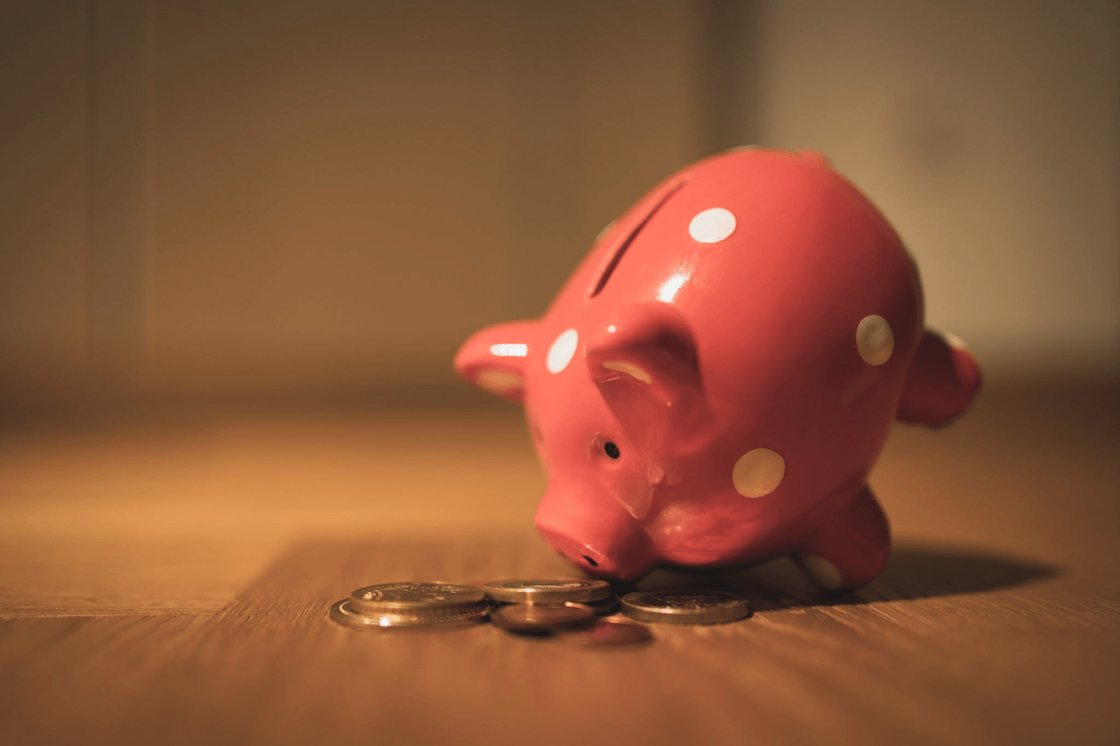 A pink piggy bank lying beside a small stack of coins on a wooden surface.